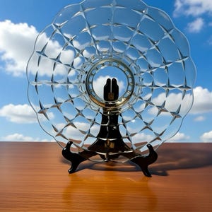 May include: A clear glass plate with a diamond pattern, displayed on a black stand. The plate has a scalloped edge and a central design. The background shows a blue sky with white clouds, creating a visually appealing display.