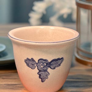 May include: A small, light pink ceramic planter with a blue floral design. The planter has a slightly flared shape and a blue rim. The design is a stylized flower with leaves. The planter is sitting on a wooden surface.