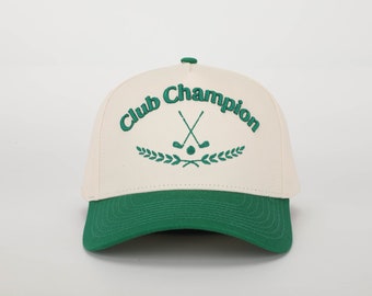 May include: A white and green baseball cap with the text "Club Champion" embroidered on the front. The cap features a green embroidered design of two crossed golf clubs and a golf ball with a wreath of green leaves.