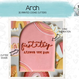 May include: A pink arch-shaped cookie cutter with the text "first trip around the sun" in red lettering. The cookie cutter is decorated with rainbow stripes in yellow, orange, and white.