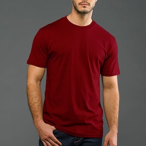 May include: A man is wearing a plain, solid red t-shirt with short sleeves. He is wearing blue jeans and has a casual, relaxed look.