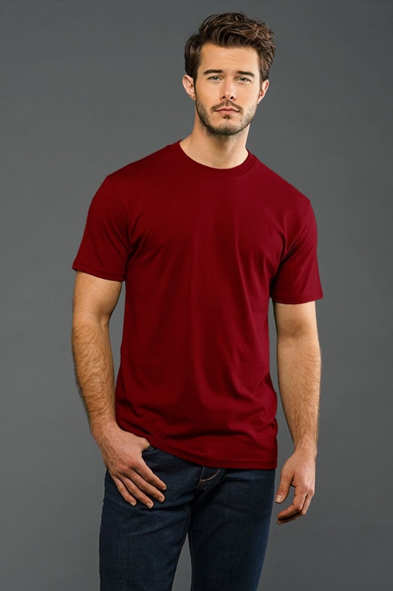 May include: A man is wearing a plain, solid red t-shirt with short sleeves. He is wearing blue jeans and has a casual, relaxed look.