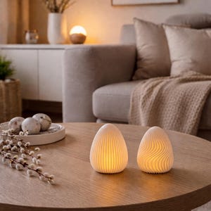 Może przedstawiać: Dwie ceramiczne lampy w kształcie jajka z ciepłym oświetleniem wewnętrznym stoją na drewnianym stoliku kawowym. Jedna lampa ma pionowe linie, druga falisty wzór. Stół znajduje się w salonie.