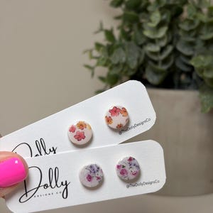 May include: Round floral stud earrings in pink, orange, and purple. The earrings are displayed on white cards with the text "Dolly Designs Co" and the Instagram handle "@TheDollyDesignsCo". The earrings have a floral pattern.