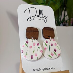May include: A pair of earrings with a white floral design of pink and purple tulips. The earrings have a wooden square top and a rounded bottom. The card behind the earrings has the text "Dolly Designs Co" and "@TheDollyDesignsCo".