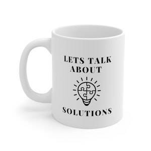 Op de afbeelding: Witte keramische mok met een zwart ontwerp met de tekst "Lets Talk About Solutions" met een lampicoon met een puzzelstuk erin.