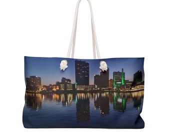 May include: A blue canvas tote bag with white rope handles. The bag features a cityscape at night with tall buildings reflected in the water.