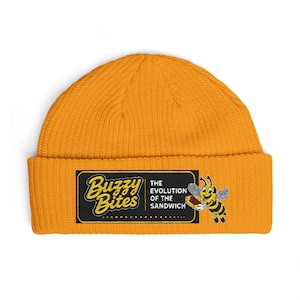 May include: Mustard yellow beanie hat with a rectangular patch featuring the text "Buzzy Bites" and a cartoon bee holding a sandwich. The patch also includes the text "The Evolution of the Sandwich".