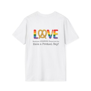 Op de afbeelding: Wit t-shirt met een kleurrijk "LOVE" ontwerp, waarbij de "O" is vervangen door een pretzel. Het woord "LOVE" is in regenboogkleuren. Onder het ontwerp staat de tekst: "Because EVERYONE Deserves to Have a Pretzel Day!"