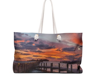 May include: A white canvas tote bag with rope handles features a colorful sunset over a wooden dock with a boat in the distance.