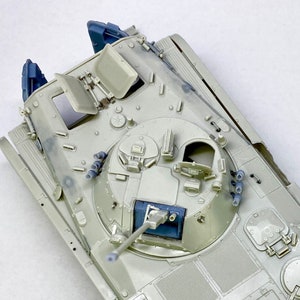 Puede incluir: Una vista cercana de un tanque modelo con un acabado de pintura gris. El tanque tiene una torreta con un cañón azul y una escotilla abierta en la parte superior. El tanque tiene un diseño detallado con varias escotillas y compartimentos.