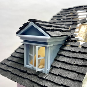 May include: A model house with a gray roof and a dormer window with a white frame. Some of the roof shingles are missing, creating a distressed look.