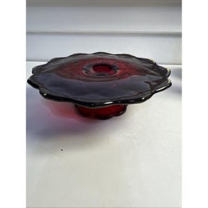 Puede incluir: Un soporte para pasteles de vidrio rojo rubí con borde festoneado. El soporte tiene un borde rojo oscuro, casi negro, y una base a juego. El centro del soporte tiene una pequeña hendidura circular. El soporte es probablemente vintage.
