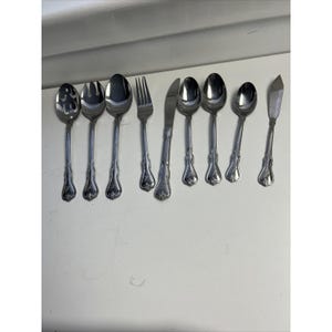 May include: A set of eight stainless steel flatware pieces arranged in a row, including a slotted spoon, a serving spoon, a dinner spoon, a dinner fork, a dinner knife, two smaller spoons, and a butter knife, all featuring a decorative handle design.