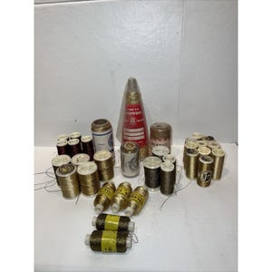 Può includere: Una collezione di rocchetti di filo da cucito metallizzato oro e bronzo di varie dimensioni e marche. L'assortimento comprende rocchetti cilindrici e un rocchetto conico avvolto in plastica trasparente, con etichette recanti testi come Lumeya, Bernat e DMC.
