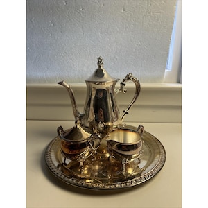 May include: A silver tea set with a teapot, creamer, sugar bowl, and serving tray. The set has ornate handles and a reflective surface. The tray is round with a decorative edge. The set is placed on a white surface.