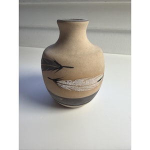May include: A beige ceramic vase with a narrow neck and a rounded body. The vase features two black and white feather designs. A dark gray band encircles the base of the vase.