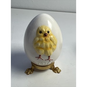 Puede incluir: Figura de huevo de cerámica blanca con diseño de pollito amarillo. El pollito tiene pico y patas rojas. El huevo se apoya en un soporte de metal dorado con patas de garra. La base del huevo dice "1976".
