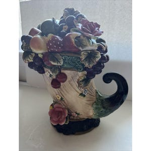 Könnte beinhalten: Eine Keramik-Schöpfungshorn mit einem Griff, verziert mit verschiedenen Früchten, Blumen und Blättern. Das Schöpfungshorn ist in Grün-, Braun-, Rot- und Violetttönen bemalt.