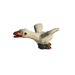 May include: A white ceramic goose figurine with outstretched wings, a bright orange beak, and a black eye. The goose is positioned as if in motion, with its head extended forward and its feet resting on a small, dark green base.