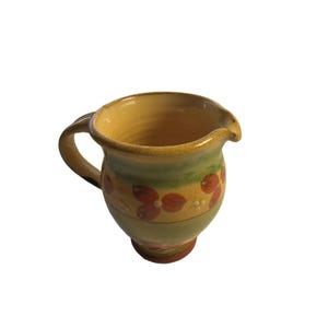 Può includere: Una brocca in ceramica con corpo arrotondato e manico curvo. L'esterno è decorato con fasce orizzontali nei toni del giallo e del verde spento, arricchite da un motivo ripetitivo di piccoli fiori rosso-arancio e puntini bianchi.
