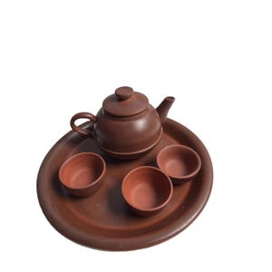 May include: A brown clay tea set consisting of a round teapot with a lid and handle, accompanied by three small matching tea cups. All items are arranged on a large, circular brown clay serving tray, featuring a smooth, matte finish throughout.