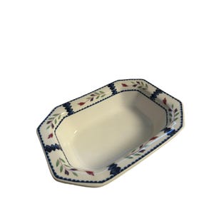 May include: An off-white ceramic serving dish with an octagonal shape. The rim features a decorative pattern of dark blue scalloped edges, interspersed with small green leaves and red floral accents painted along the border of the bowl.