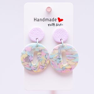 Könnte beinhalten: Handgefertigte Ohrringe in Pastelltönen mit Marmoreffekt. Jeder Ohrring hat einen strukturierten, hellvioletten Kreis über einem größeren, offen zentrierten Kreis mit einer Mischung aus Pastellfarben. Auf der Karte steht "Handmade with love".