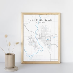 Puede incluir: Una impresión enmarcada del mapa de Lethbridge, Alberta, en un estilo minimalista. El mapa presenta un patrón de cuadrícula con líneas negras y un río azul claro. El nombre de la ciudad se muestra en la parte superior. El marco es de madera clara.