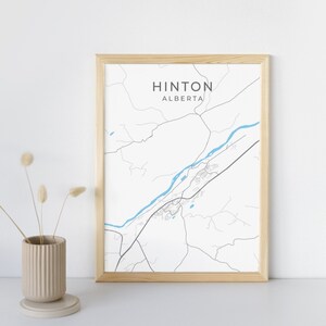 Op de afbeelding: Een ingelijste print met een minimalistische kaart van Hinton, Alberta. De kaart is in zwart en wit met een blauwe rivier en de stadsnaam bovenaan. De lijst is van licht hout en links staat een vaas met gedroogde bloemen.