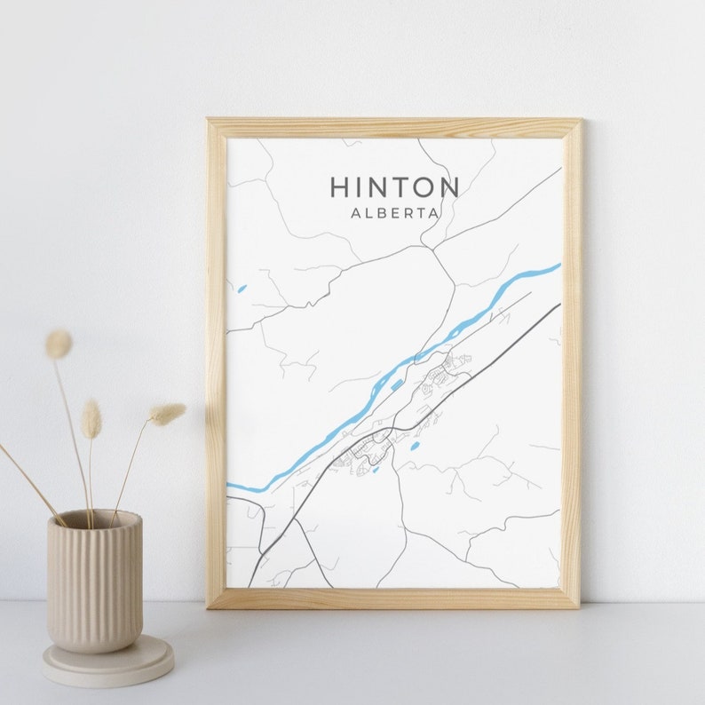 Op de afbeelding: Een ingelijste print met een minimalistische kaart van Hinton, Alberta. De kaart is in zwart en wit met een blauwe rivier en de stadsnaam bovenaan. De lijst is van licht hout en links staat een vaas met gedroogde bloemen.