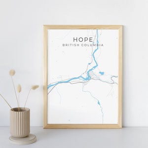 May include: A framed map print of Hope, British Columbia, with a light blue river and road design on a white background. The print is in a light wood frame and is displayed on a white surface.