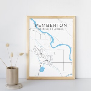 Peut inclure: Une impression encadrée d'une carte de Pemberton, en Colombie-Britannique, avec des détails bleus clairs pour la rivière et le lac. La carte est dans un cadre en bois clair et présente le nom de la ville dans une police moderne. Un vase beige avec des fleurs séchées est à gauche.