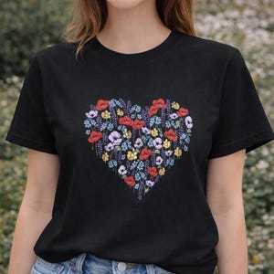 Könnte beinhalten: Schwarzes T-Shirt mit einem herzförmigen Blumendesign. Das Herz besteht aus roten Mohnblumen, lila, gelben und blauen Blumen. Das T-Shirt hat einen Rundhalsausschnitt.