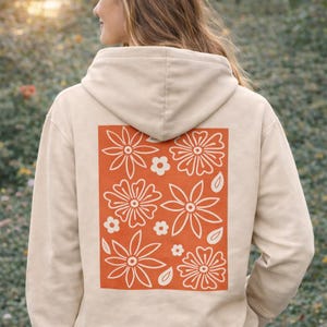 Könnte beinhalten: Beigefarbener Kapuzenpullover mit floralem Design auf dem Rücken. Das Design ist ein rechteckiger orangefarbener Block mit weiß umrandeten Blumen und Blättern. Der Kapuzenpullover hat eine Kapuze und ist aus weichem Material.