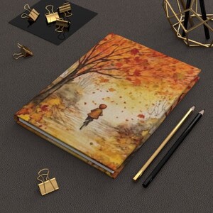 May include: A watercolor painting of a person walking through a forest with autumn leaves falling. The painting is on the cover of a journal.