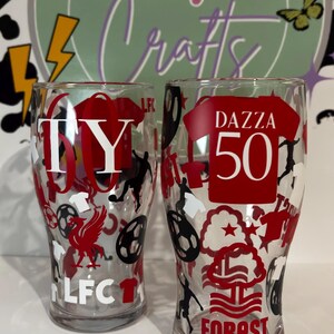 May include: Two clear pint glasses with red and black soccer-themed designs. One glass has "TY 60" and "LFC" text, the other has "DAZZA 50" and "FOREST" text. The glasses are decorated with soccer balls, players, and team logos.
