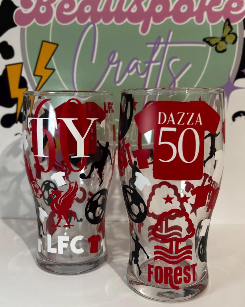May include: Two clear pint glasses with red and black soccer-themed designs. One glass has "TY 60" and "LFC" text, the other has "DAZZA 50" and "FOREST" text. The glasses are decorated with soccer balls, players, and team logos.