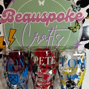 May include: Three clear pint glasses with personalized names and soccer team logos. The glasses feature the names "JON", "PETE", and "TOM" in various colors, along with team emblems and the text "AFC". The background includes a logo that says "Beauspoke Crafts".
