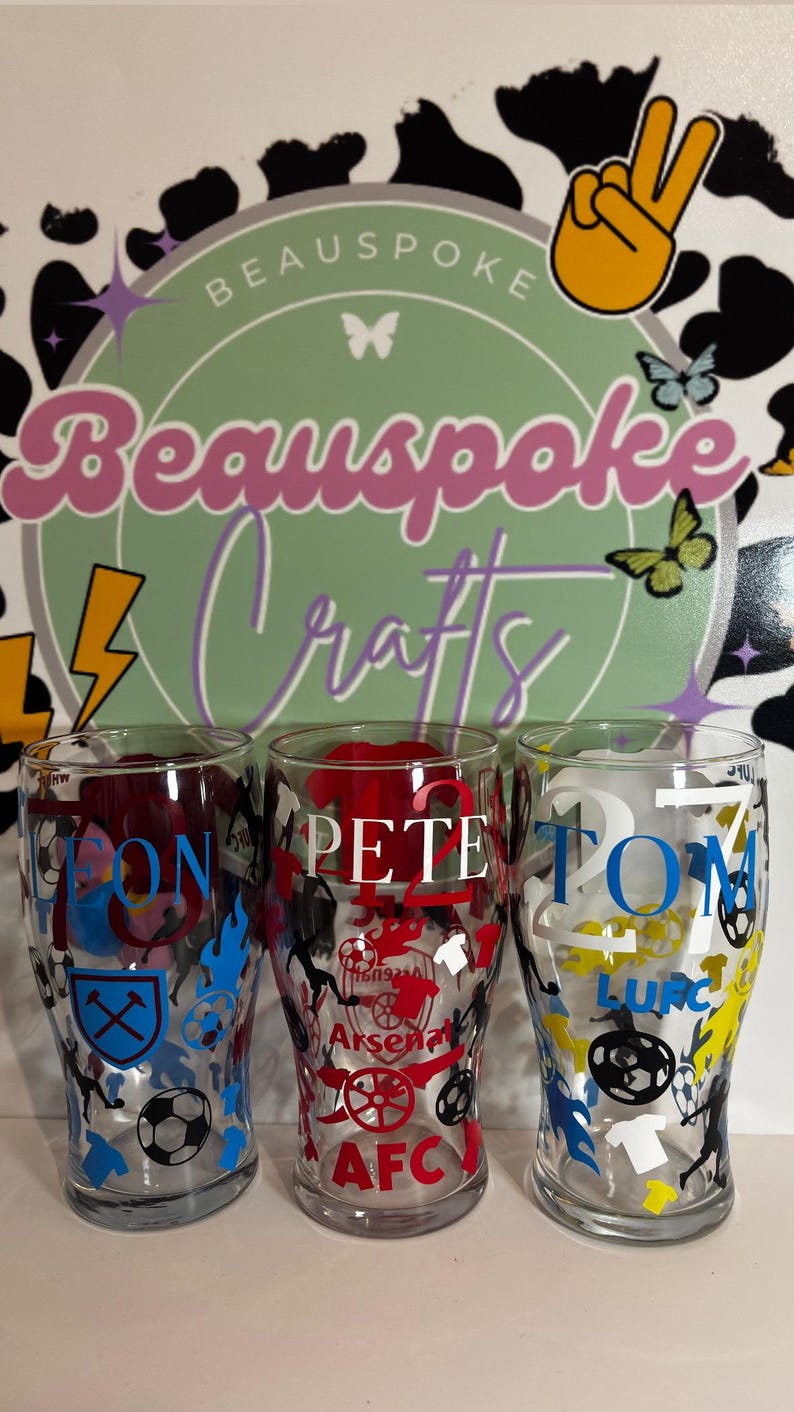 May include: Three clear pint glasses with personalized names and soccer team logos. The glasses feature the names "JON", "PETE", and "TOM" in various colors, along with team emblems and the text "AFC". The background includes a logo that says "Beauspoke Crafts".