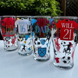 May include: Four clear pint glasses with soccer-themed designs. Each glass features a jersey design with names and numbers, along with soccer balls, player silhouettes, and team logos. The text includes "DAD", "WILL 21", "BRANDON 16", and "ADD 22".