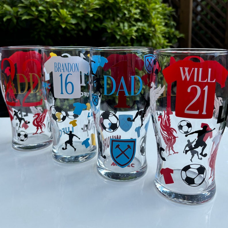 May include: Four clear pint glasses with soccer-themed designs. Each glass features a jersey design with names and numbers, along with soccer balls, player silhouettes, and team logos. The text includes "DAD", "WILL 21", "BRANDON 16", and "ADD 22".