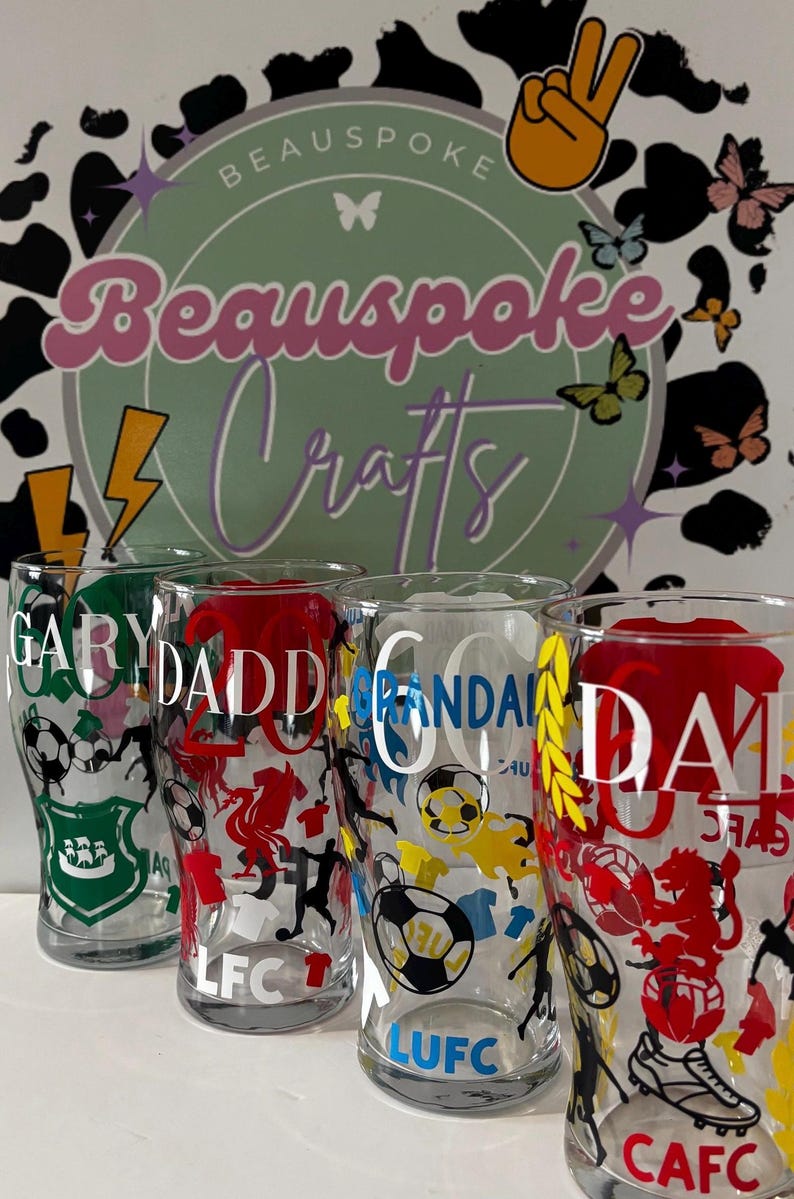 May include: Four clear pint glasses with custom designs. Each glass features a name and team-related graphics in various colors, including green, red, blue, and yellow. The background includes a logo with the text "Beauspoke Crafts" and decorative butterflies.