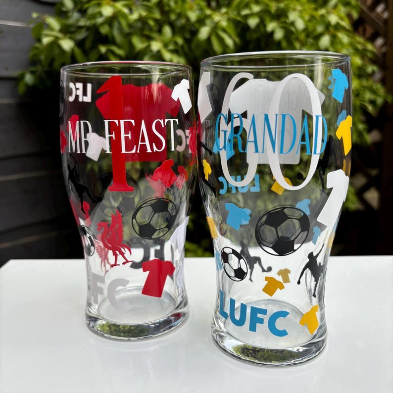 May include: Two clear pint glasses with soccer-themed designs. One glass has "MR FEAST" in white text with red accents and a soccer ball graphic. The other glass has "GRANDAD" in white text with blue accents and a soccer ball graphic.