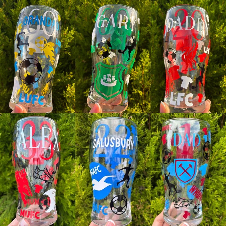 May include: Six clear pint glasses with soccer-themed designs. Each glass features a name, number, and soccer-related graphics in various colors, including red, green, blue, and yellow. The glasses are displayed against a backdrop of green foliage.