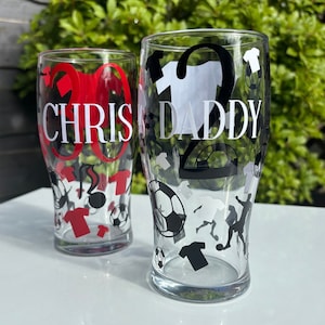 May include: Two clear pint glasses with soccer-themed designs. One glass has "CHRIS" and the number "30" in red. The other glass has "DADDY" and the number "2" in black. Both glasses feature soccer balls and jerseys.