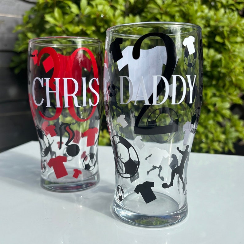 May include: Two clear pint glasses with soccer-themed designs. One glass has "CHRIS" and the number "30" in red. The other glass has "DADDY" and the number "2" in black. Both glasses feature soccer balls and jerseys.