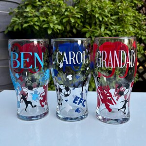 May include: Three clear pint glasses with personalized text and soccer-themed designs. The glasses feature names in blue and red, with soccer ball and player graphics. One glass has "EFC" text.
