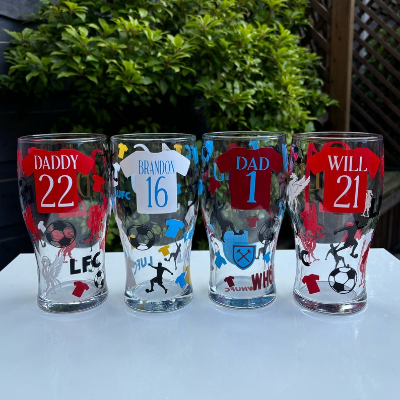 May include: Four clear pint glasses with soccer-themed designs. Each glass features a jersey design with a name and number, along with soccer ball and player graphics. The text includes "DADDY 22", "BRANDON 16", "DAD 1", and "WILL 21".