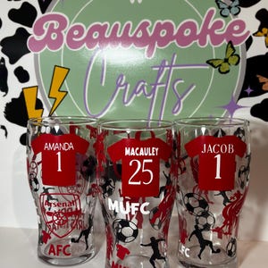 May include: Three clear pint glasses with red soccer jersey designs. Each glass features a name and number, with team logos and soccer ball graphics. The names are Amanda, Macauley, and Jacob. The background includes a logo that says "Beauspoke Crafts".